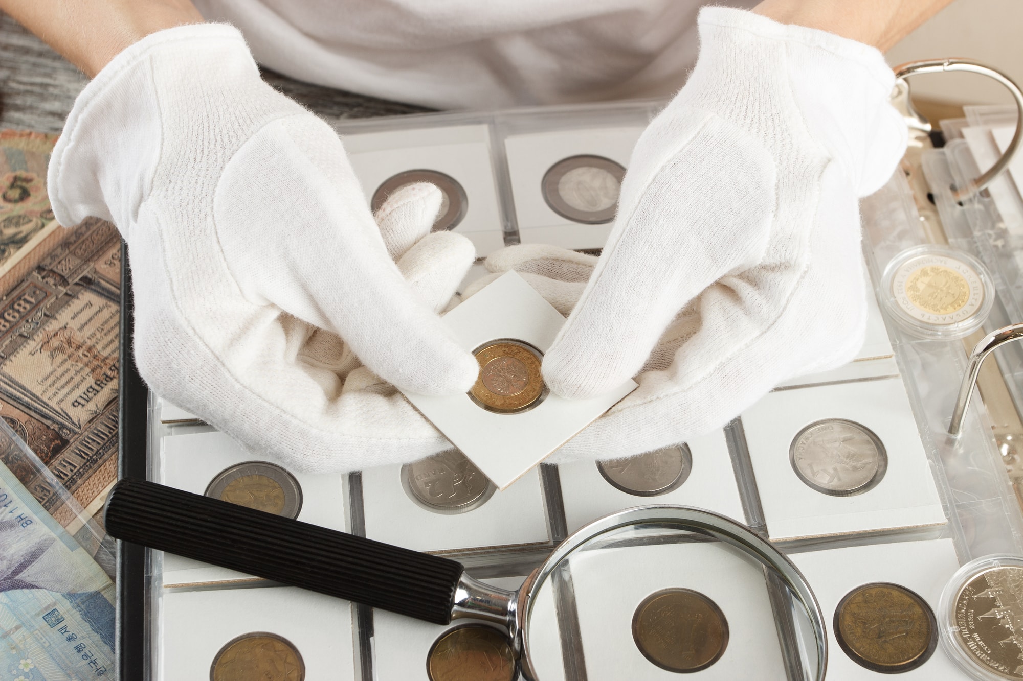 A person wearing white gloves uses a magnifying glass to inspect a box filled with various coins, highlighting their features.